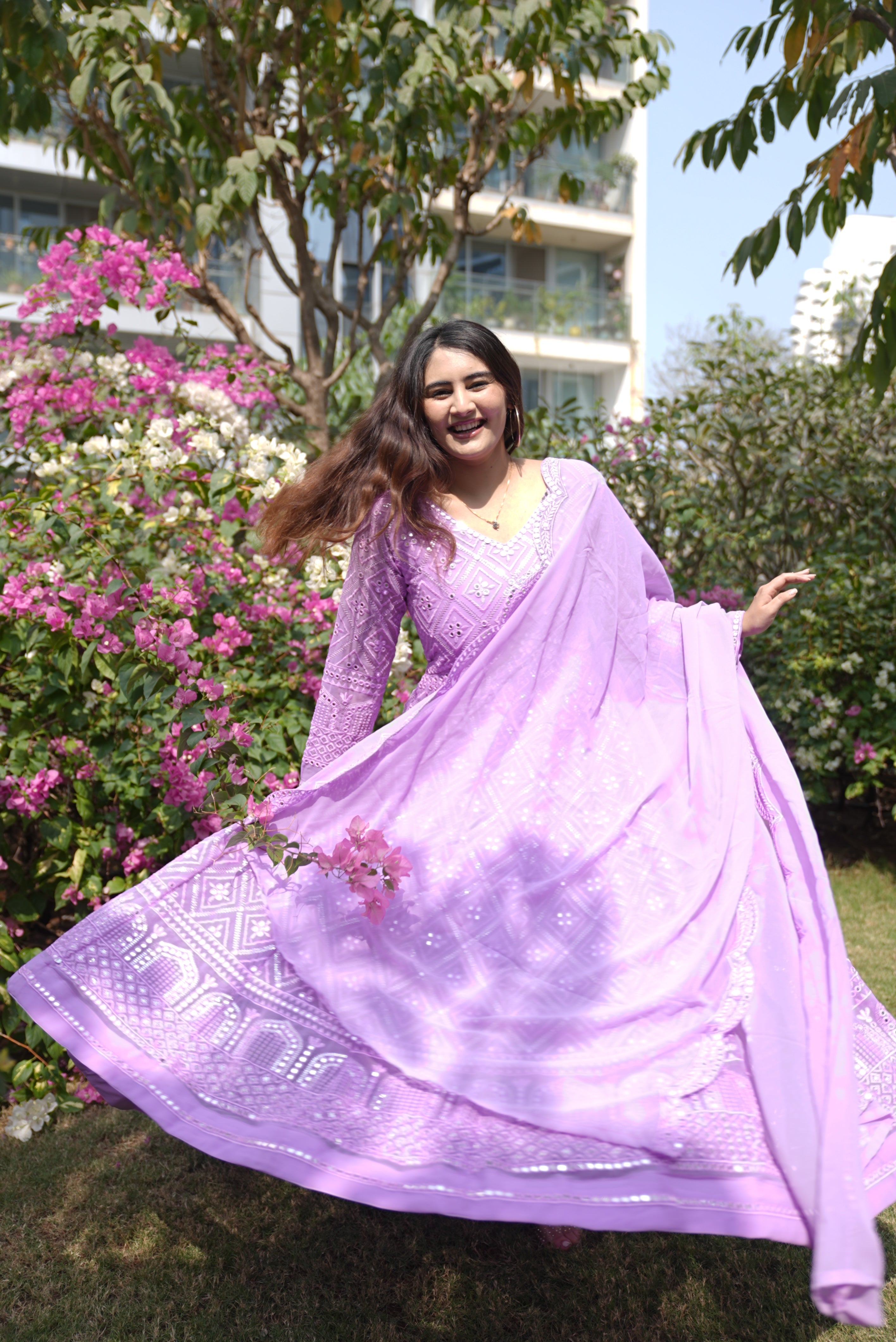 Woman in a purple dress standing outdoors with greenery and flowers in the background